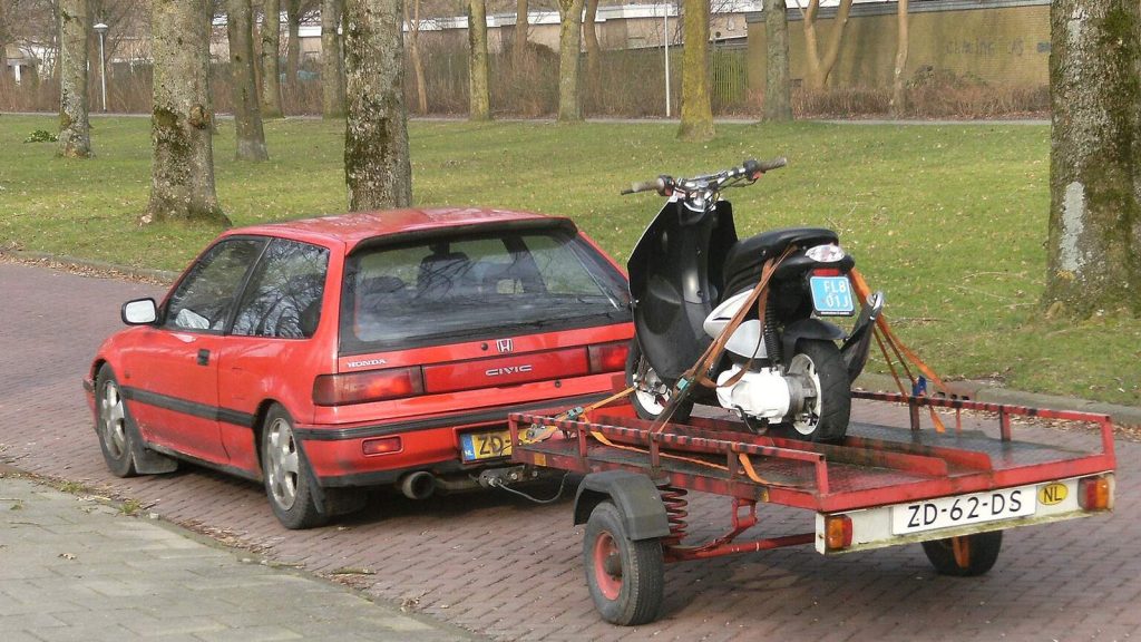 Voiture rouge tractant une remorque plateau avec un scooter sanglé dessus
